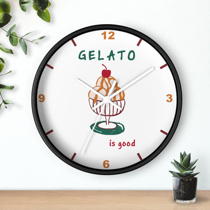 A round wall clock with a black frame and a white face, featuring a gelato cupcake illustration and the words 'GELATO is good'. The clock is mounted on a white wall above a light wood surface, with decorative green leaves and a small succulent nearby.