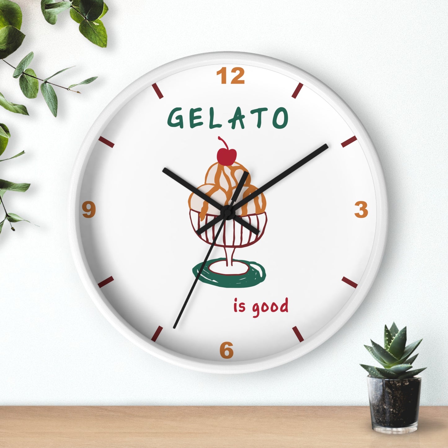 A round white wall clock with a colorful gelato cupcake illustration in the center, brown and orange numerals, and the words 'GELATO is good' on the face. The clock is mounted on a white wall above a light wood surface, with small green plants in the corners.
