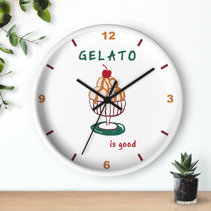 A round white wall clock with a colorful gelato cupcake illustration in the center, brown and orange numerals, and the words 'GELATO is good' on the face. The clock is mounted on a white wall above a light wood surface, with small green plants in the corners.