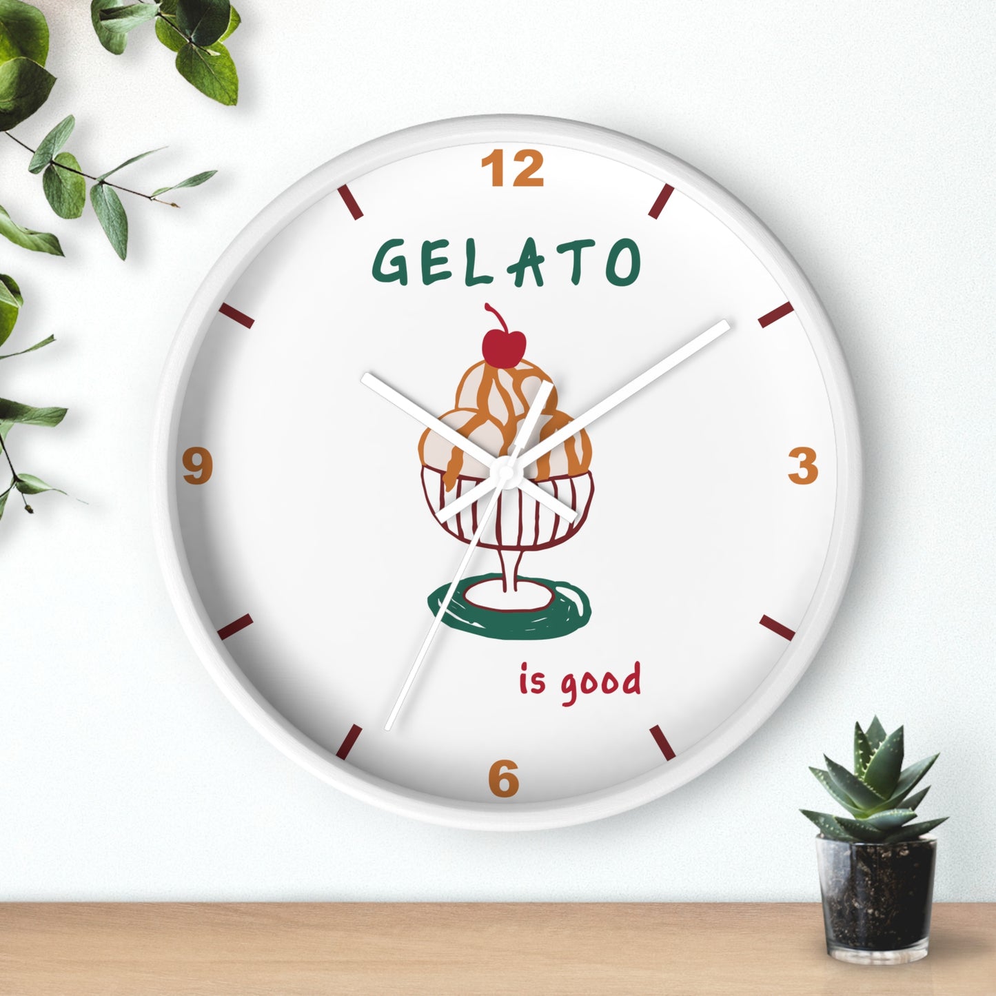 A round white wall clock with a colorful gelato cupcake illustration in the center, featuring the text 'GELATO is good' and brown hour markers, styled on a light wall above a wooden surface with small green plants.