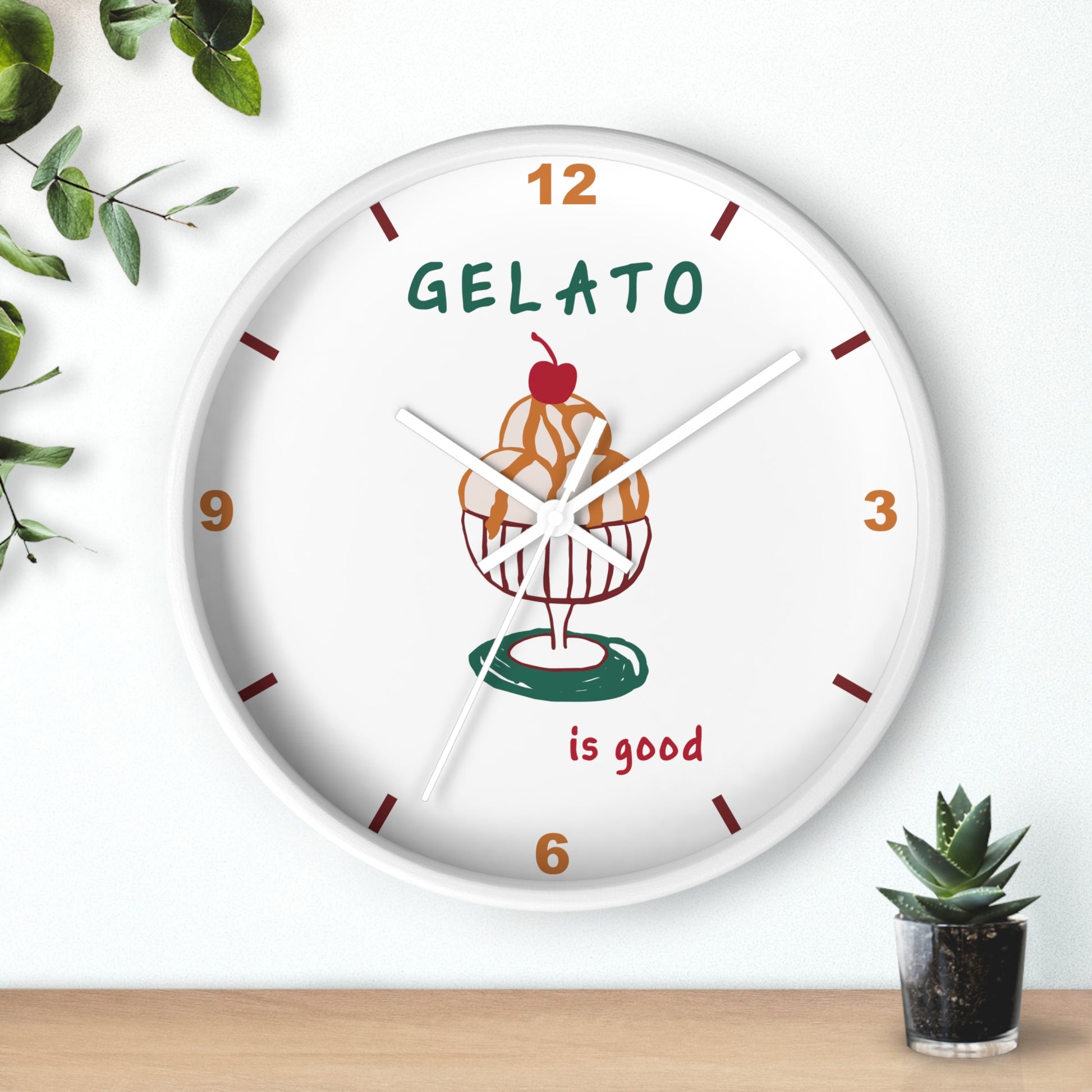A round white wall clock with a colorful gelato cupcake illustration in the center, featuring the text 'GELATO is good' and brown hour markers, styled on a light wall above a wooden surface with small green plants.