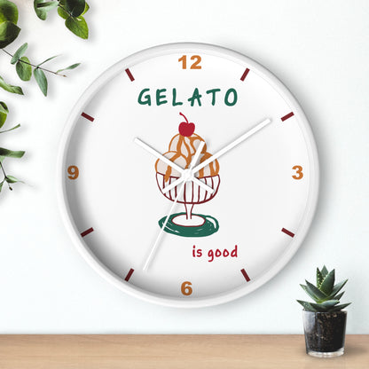 A round white wall clock with a colorful gelato cupcake illustration in the center, featuring the text 'GELATO is good' and brown hour markers, styled on a light wall above a wooden surface with small green plants.