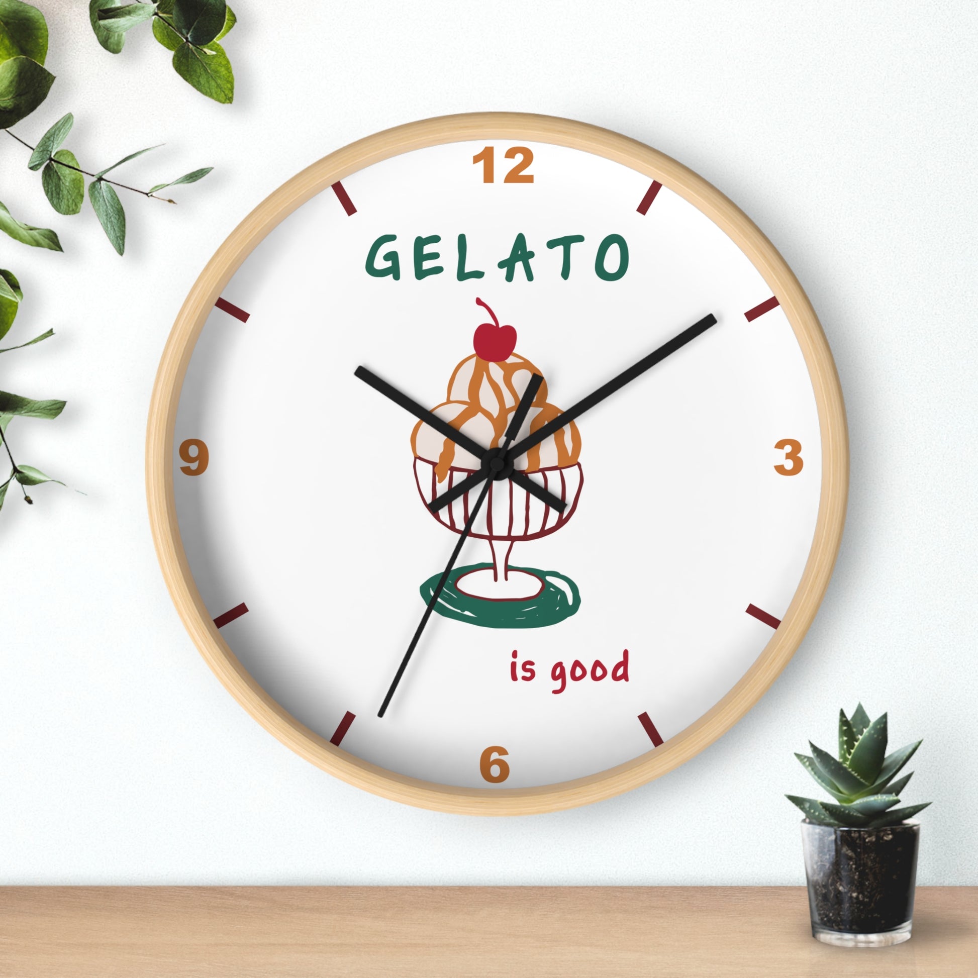 A round wall clock with a light wood frame and a white face, featuring a gelato cupcake illustration and the words 'GELATO is good'. The clock is mounted on a white wall above a wooden surface, with decorative green leaves and a small potted succulent nearby.