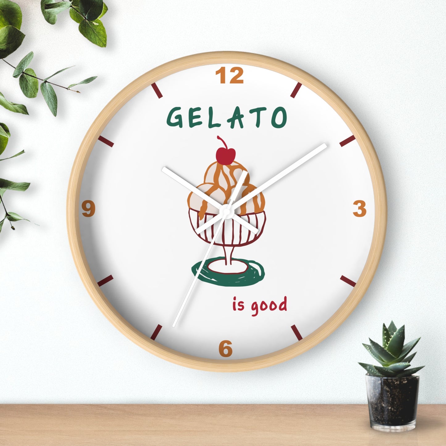 A round wall clock with a light wood frame and a white face, featuring a gelato cup illustration and the words 'GELATO is good'. The clock is styled on a wooden surface against a white wall with decorative green leaves and a small potted succulent.