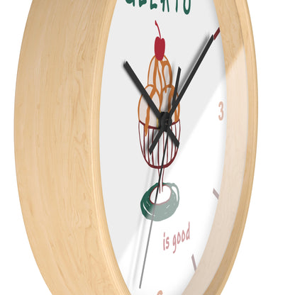 A close-up of a round wall clock with a light natural wood frame and a white dial featuring a colorful ice cream cone illustration and the words 'GELATO is good'.