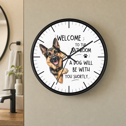 A round wall clock with a black frame and a white face, featuring a German Shepherd illustration and the text 'WELCOME TO THE BATHROOM A DOG WILL BE WITH YOU SHORTLY.' The clock is mounted on a beige wall in a bathroom setting.