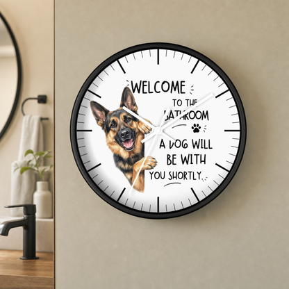 A round wall clock with a black frame and a white face, featuring a German Shepherd illustration and the text 'WELCOME TO THE BATHROOM A DOG WILL BE WITH YOU SHORTLY.' The clock is mounted on a beige wall in a bathroom setting.