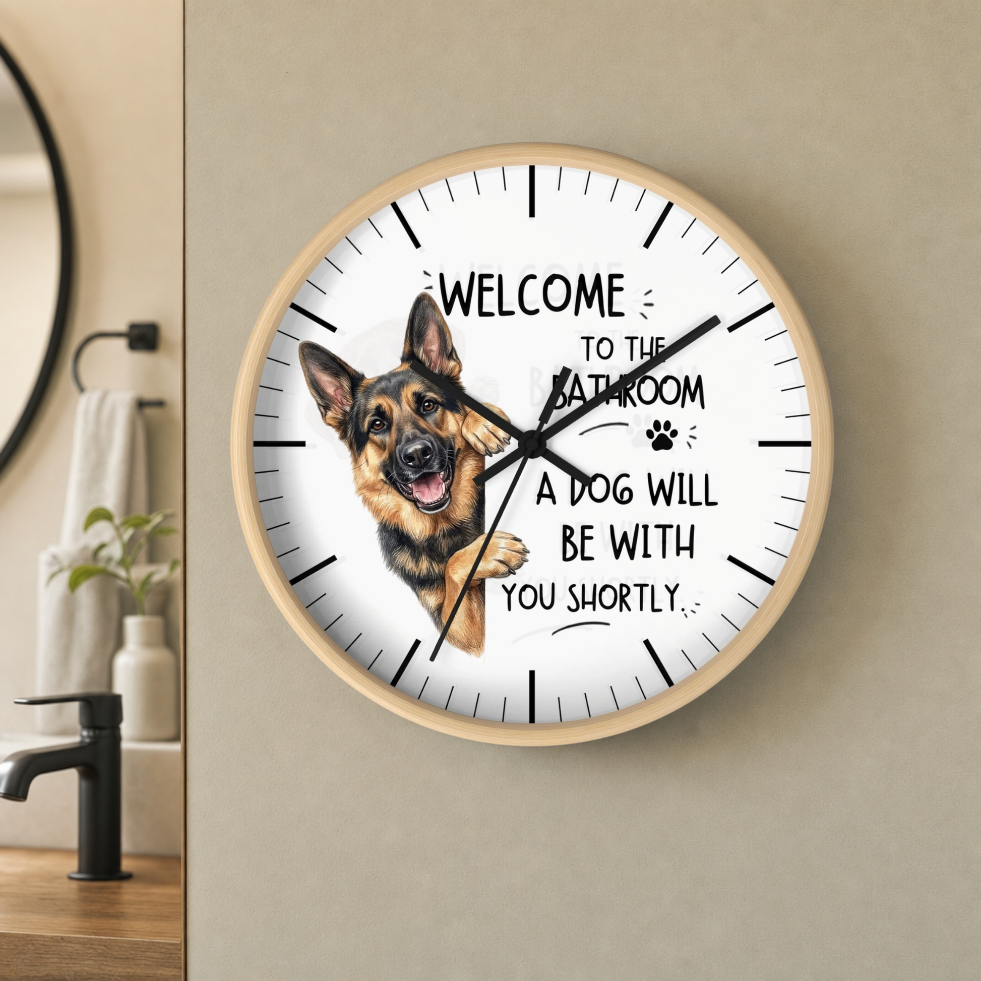 A round wall clock with a light wood frame and a white face, featuring a German Shepherd illustration and the text 'WELCOME TO THE BATHROOM A DOG WILL BE WITH YOU SHORTLY.' The clock is mounted on a beige wall in a bathroom setting.
