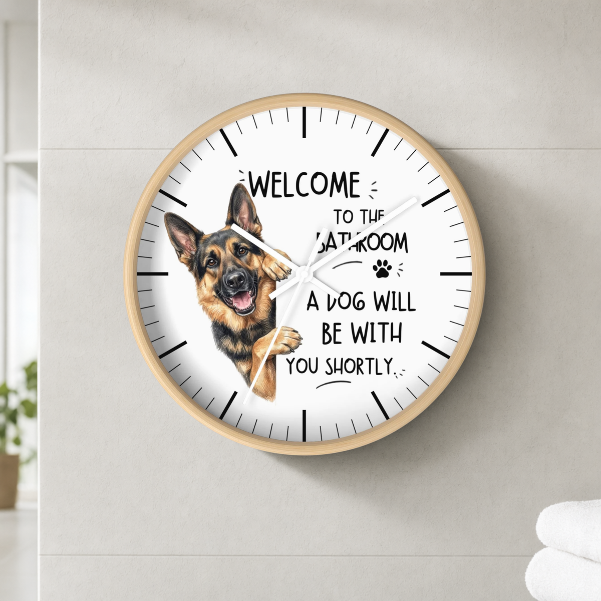 A round wall clock with a light wood frame and a white face, mounted on a light gray wall. The clock features a German Shepherd illustration and the text 'WELCOME TO THE BATH ROOM A DOG WILL BE WITH YOU SHORTLY'.