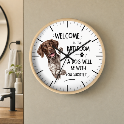 A round wall clock with a light wood frame and a white face, featuring a brown German Shorthaired Pointer dog illustration and the text 'WELCOME TO THE BATHROOM A DOG WILL BE WITH YOU SHORTLY.' The clock is mounted on a beige wall in a bathroom setting.