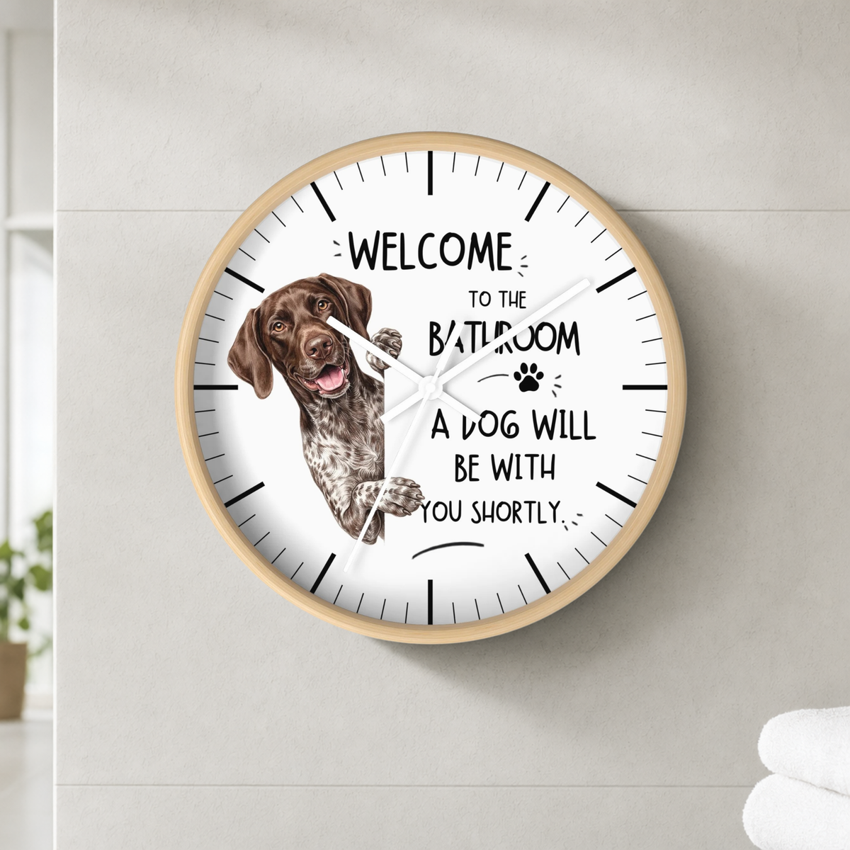 A round wall clock with a light wood frame and white face, mounted on a light gray tiled wall. The clock features a brown German Shorthaired Pointer dog illustration and the humorous text 'WELCOME TO THE BATHROOM A DOG WILL BE WITH YOU SHORTLY'.