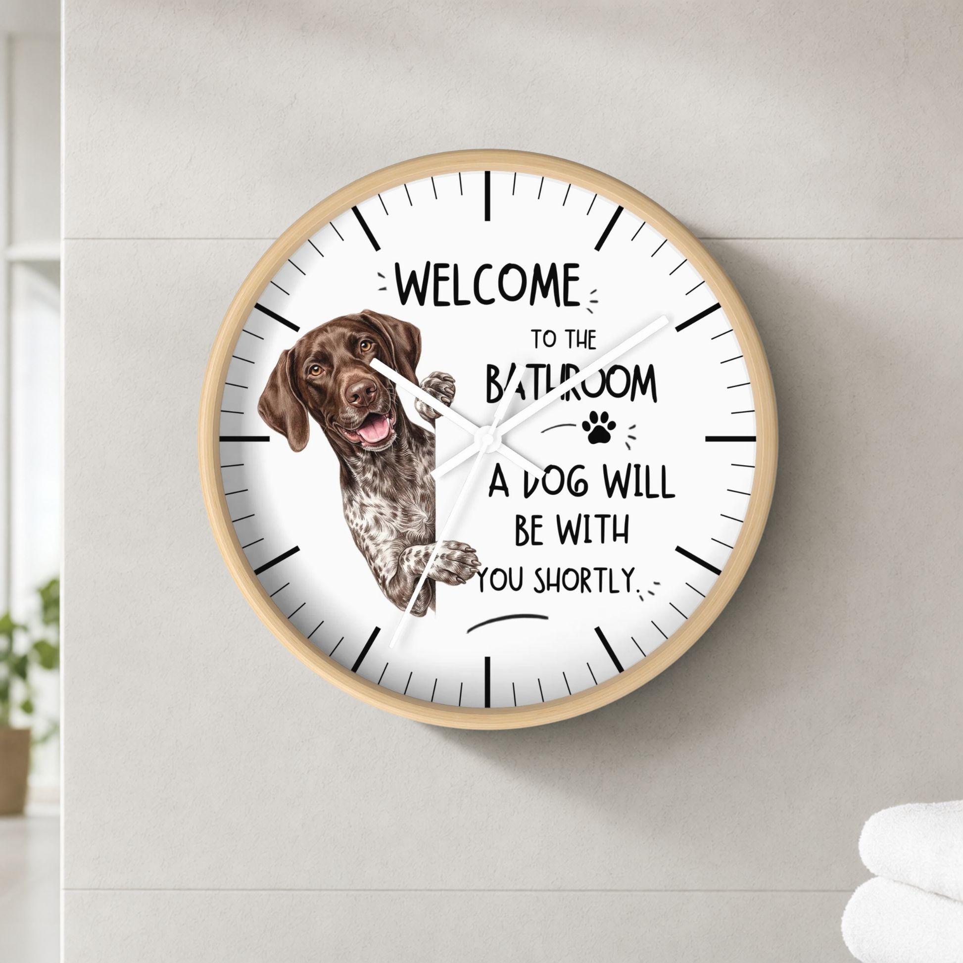 A round wall clock with a light wood frame and white face, mounted on a light gray tiled wall. The clock features a brown German Shorthaired Pointer dog illustration and the humorous text 'WELCOME TO THE BATHROOM A DOG WILL BE WITH YOU SHORTLY'.