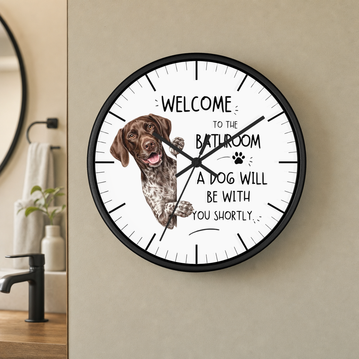 A round wall clock with a black frame and white face, mounted on a beige wall in a bathroom setting. The clock face features a brown and white spotted dog illustration and the text 'WELCOME TO THE BATHROOM A DOG WILL BE WITH YOU SHORTLY'.