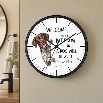 A round wall clock with a black frame and white face, mounted on a beige wall in a bathroom setting. The clock face features a brown and white spotted dog illustration and the text 'WELCOME TO THE BATHROOM A DOG WILL BE WITH YOU SHORTLY'.