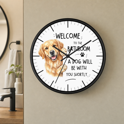 A round wall clock with a black frame and a white face, featuring a golden retriever illustration and the text 'WELCOME TO THE BATHROOM A DOG WILL BE WITH YOU SHORTLY'. The clock is mounted on a beige wall in a bathroom setting.
