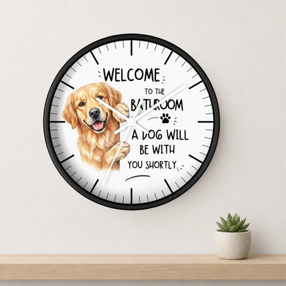 A round wall clock with a black frame and a white face, featuring a golden retriever illustration and the text 'WELCOME TO THE BATHROOM A DOG WILL BE WITH YOU SHORTLY.' The clock is mounted on a light beige wall above a wooden shelf with a small potted succulent.