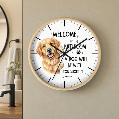 A round wall clock with a light wood frame hangs on a beige wall in a bathroom setting. The clock face is white with black text reading 'WELCOME TO THE BATHROOM A DOG WILL BE WITH YOU SHORTLY' and a large golden retriever illustration replacing the center hour marker.