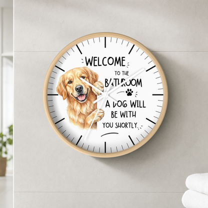 A round wall clock with a light wood frame and a white face, featuring a golden retriever illustration and the text 'WELCOME TO THE BATHROOM A DOG WILL BE WITH YOU SHORTLY'. The clock is mounted on a light gray tiled wall.