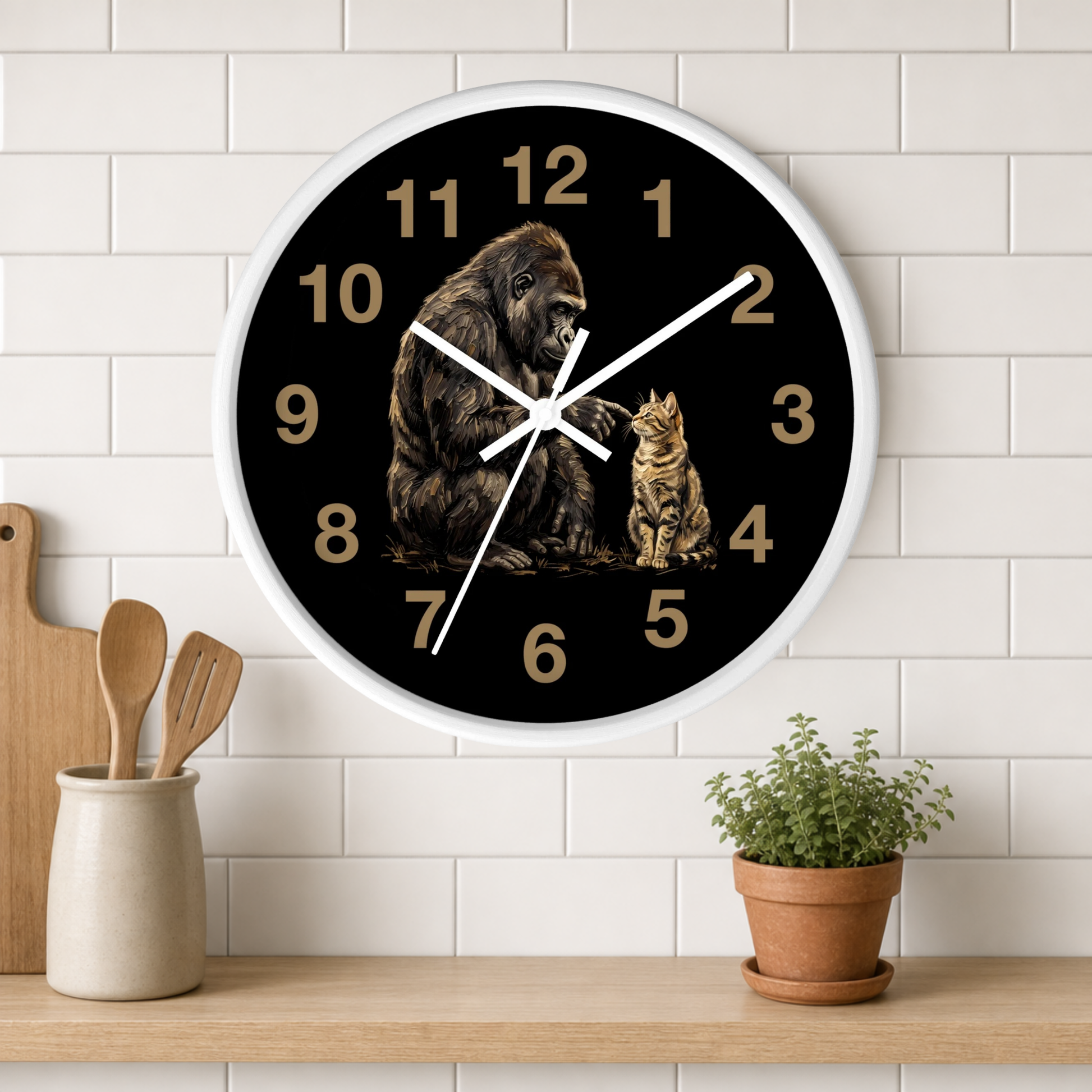 A round wall clock with a black face, white frame, and gold Arabic numerals, featuring an illustration of a gorilla and a cat. The clock is mounted on a white subway tile wall above a wooden shelf with a small potted plant and wooden utensils.