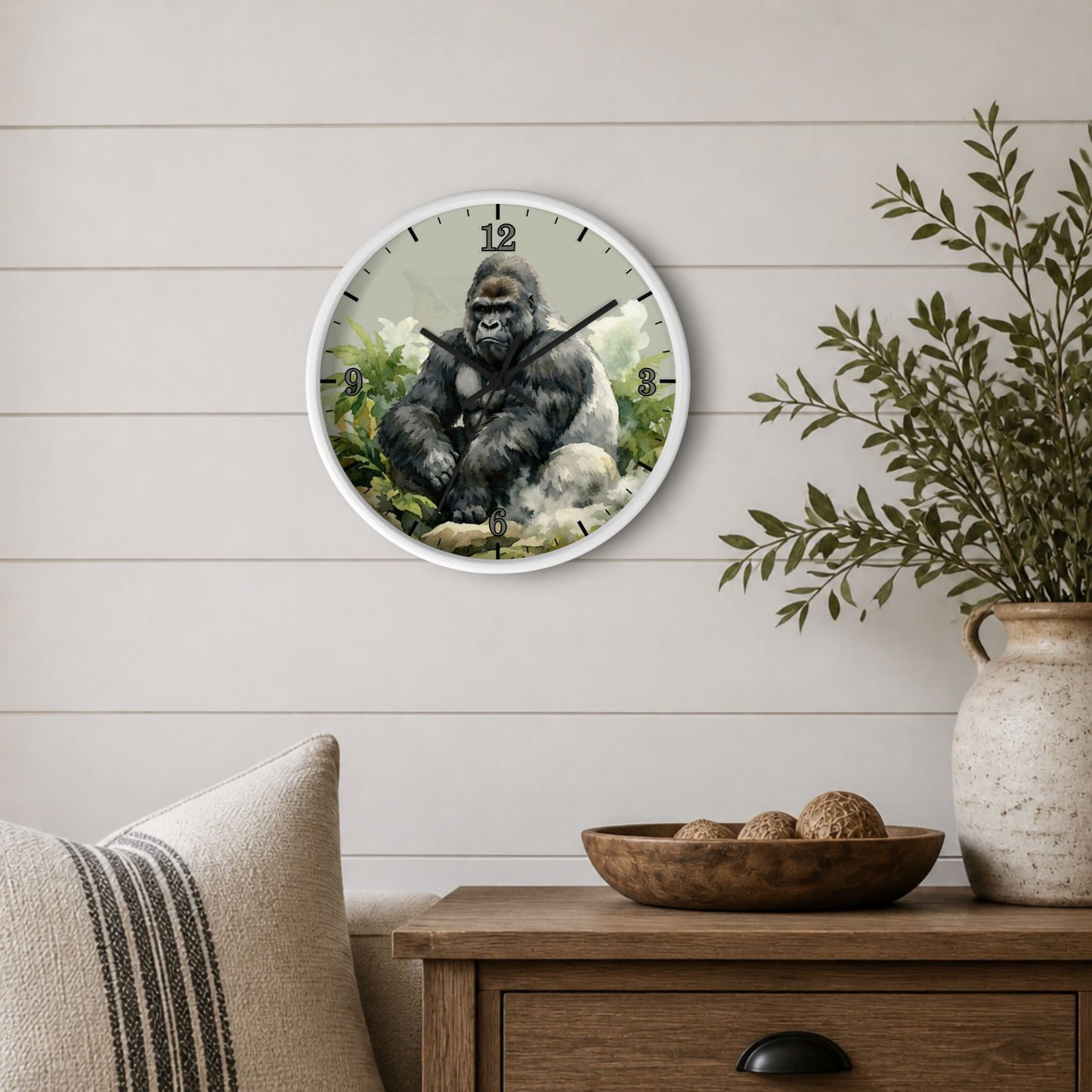 A round wall clock featuring a realistic gorilla illustration on the face, mounted on a light beige shiplap wall above a wooden side table with a vase and bowl.