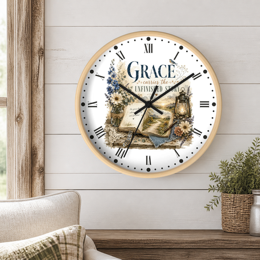 A round wall clock with a light wood frame hangs on a white shiplap wall in a styled room. The clock face is white with black Roman numerals and hands, decorated with a vintage illustration of an open book, flowers, a lantern, and the words 'GRACE carries the unfinished story'.