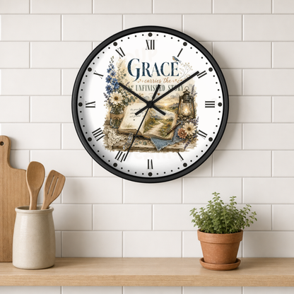 A round wall clock with a black frame and a white face featuring the word 'GRACE' and a vintage-style illustration of an open book, flowers, and a lantern. The clock is mounted on a white subway tile wall above a wooden shelf with kitchen utensils and a small potted plant.