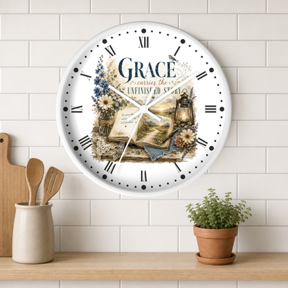 A round white wall clock with Roman numerals and a vintage-style floral illustration featuring the word 'Grace' and an open book. The clock is mounted on a white subway tile wall above a wooden shelf with a small potted plant and kitchen utensils.