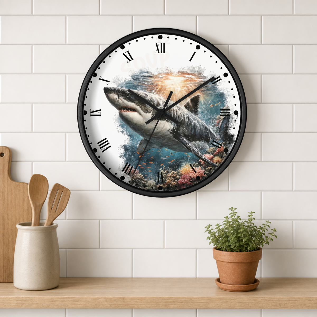 A round wall clock with a black frame and a shark underwater illustration on the face, mounted on a white subway tile wall above a wooden shelf with kitchen utensils and a potted plant.