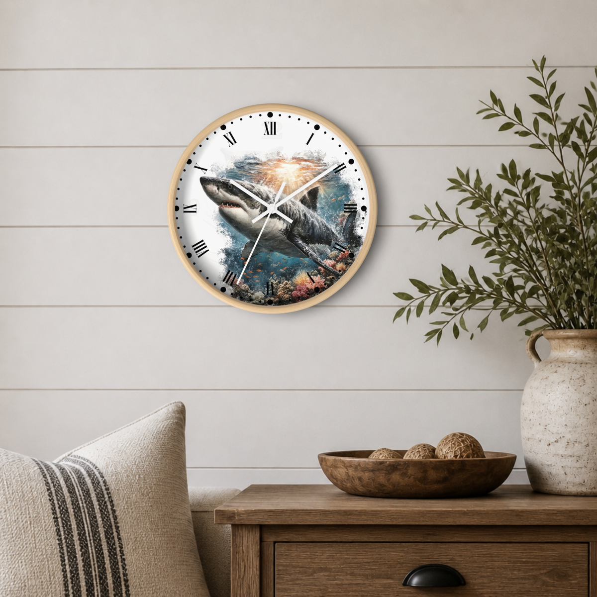 Round wall clock with a shark and ocean scene printed on the face, mounted on a white shiplap wall above a wooden side table with a vase and bowl.