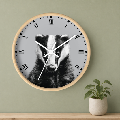 A round wall clock with a light wood frame and a gray face featuring a black-and-white badger illustration, mounted on a sage green wall above a wooden shelf with a small potted plant.