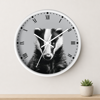 A round wall clock with a white frame and a black-and-white badger face printed on the dial, mounted on a light beige wall above a wooden surface with a small potted succulent.