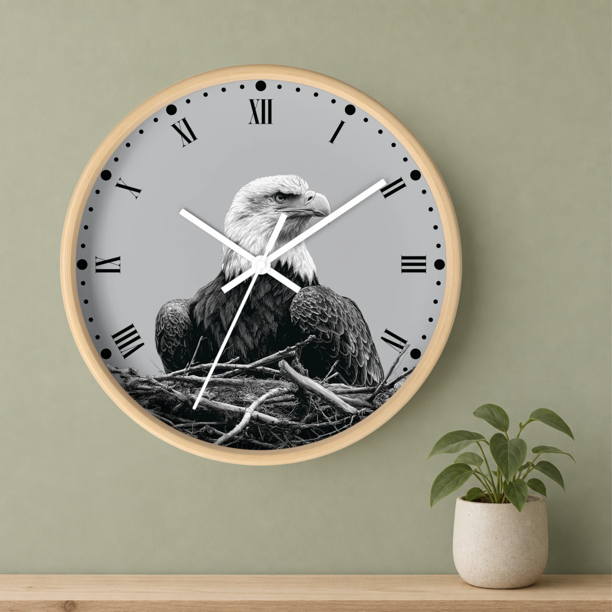 A round wall clock with a light wood frame and a grayscale eagle perched in a nest design on the face, mounted on a sage green wall above a wooden shelf with a small potted plant.
