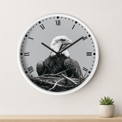A round wall clock with a white frame and Roman numerals, featuring a black-and-white photograph of a bald eagle sitting in a nest on the clock face. The clock is mounted on a light beige wall above a wooden surface with a small potted succulent.