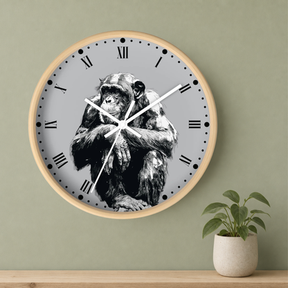 A round wall clock with a light wood frame and a gray face featuring a black-and-white illustration of a chimpanzee. The clock is mounted on a sage green wall above a wooden shelf, with a small potted plant to the right.