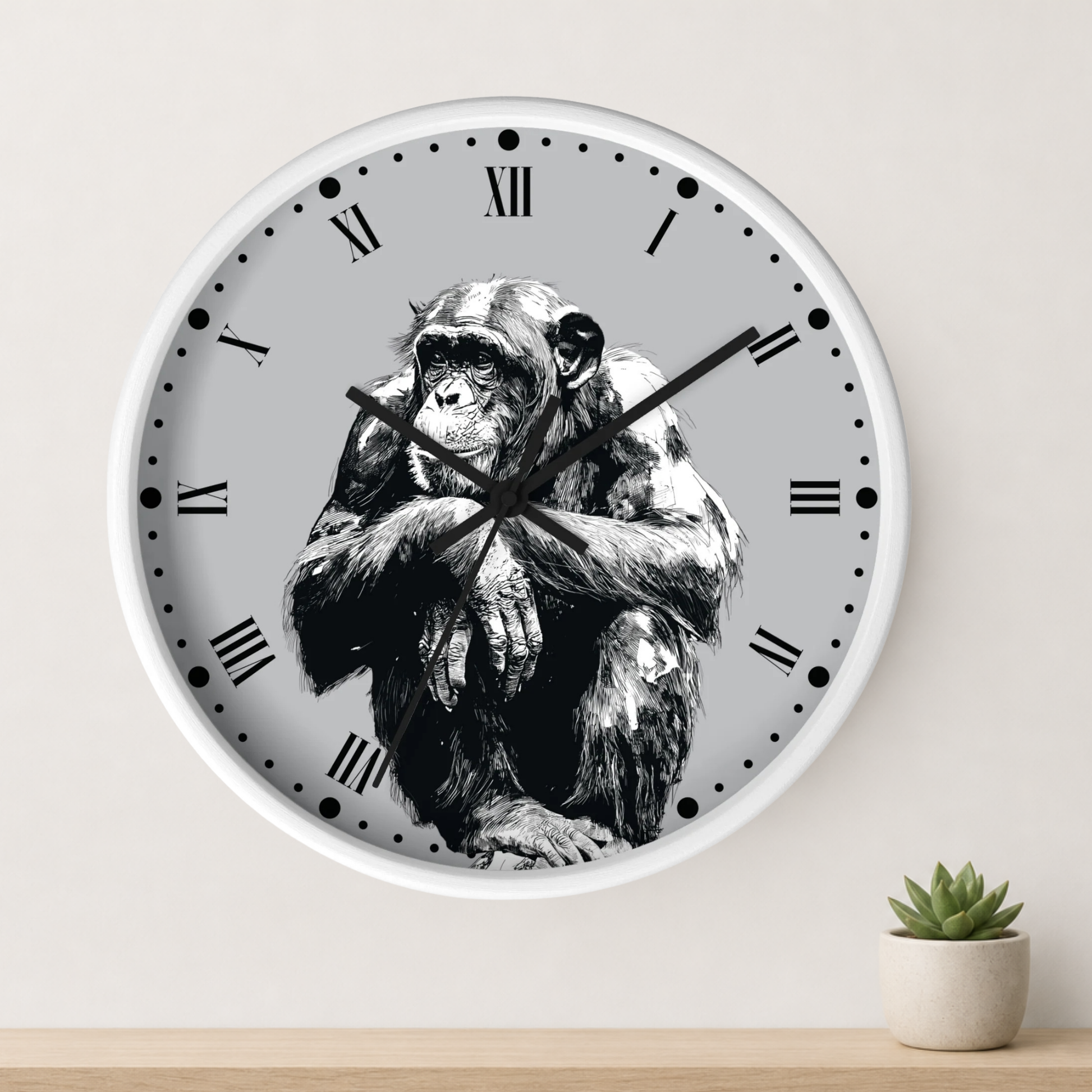 A round wall clock with a white frame and a light gray face featuring a black-and-white illustration of a seated chimpanzee. The clock is mounted on a light wall above a wooden surface, with a small potted succulent to the right.