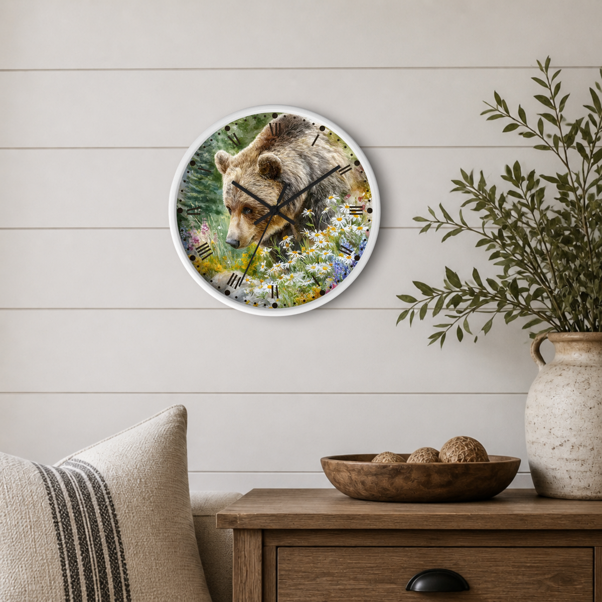 Round wall clock with a white frame featuring a bear and wildflower print, styled on a light wood paneled wall above a wooden side table with a plant and decorative bowl.