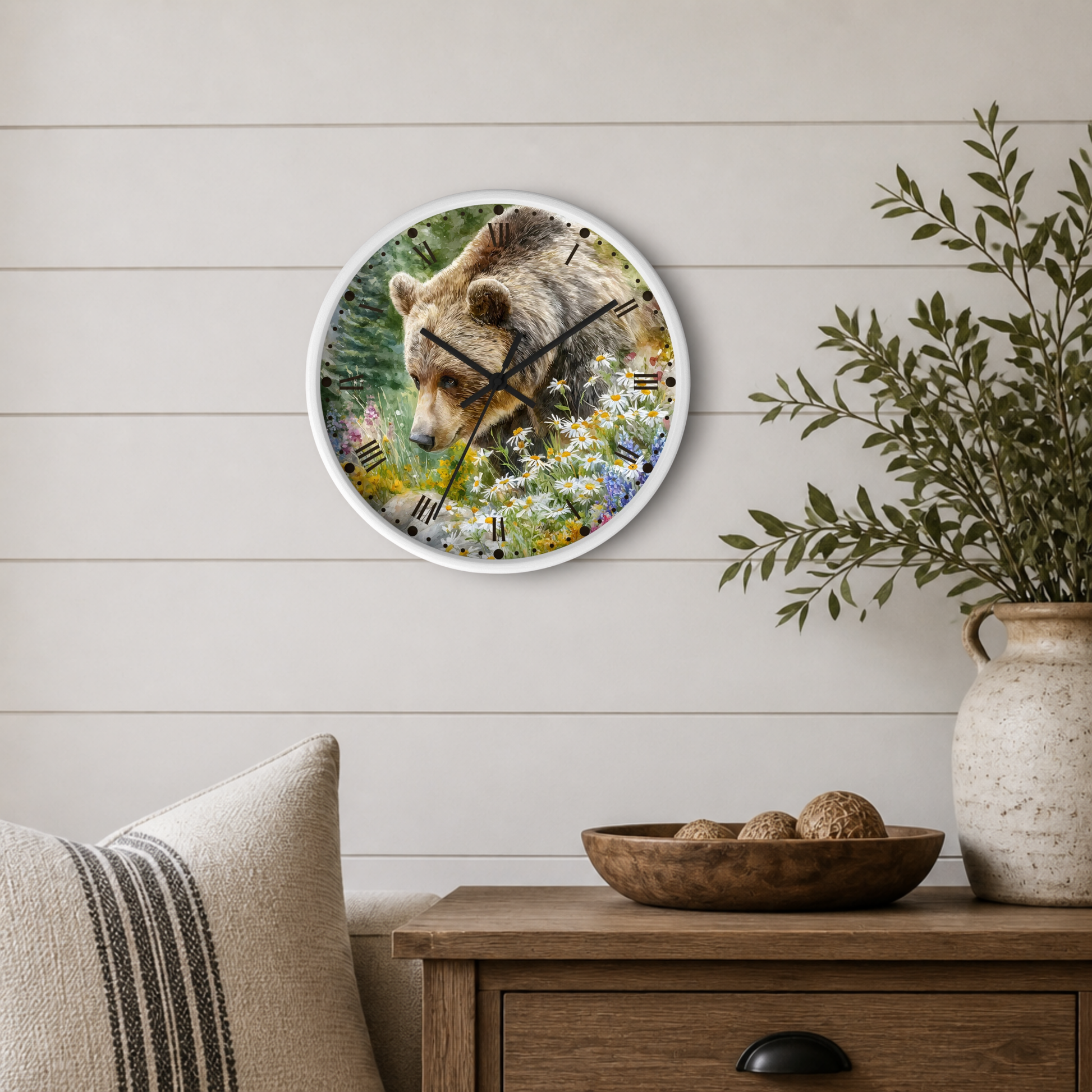 Round wall clock with a white frame featuring a bear and wildflower print, styled on a light wood paneled wall above a wooden side table with a plant and decorative bowl.