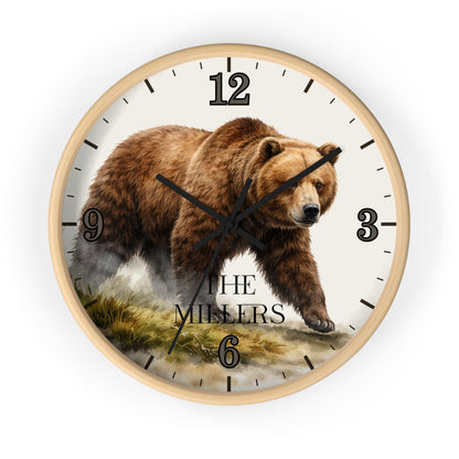 A round wall clock with a light wood frame and a printed design featuring a brown grizzly bear walking across a grassy landscape. The clock face is off-white with black Arabic numerals and hands, and the words 'THE MILLERS' printed near the bottom.