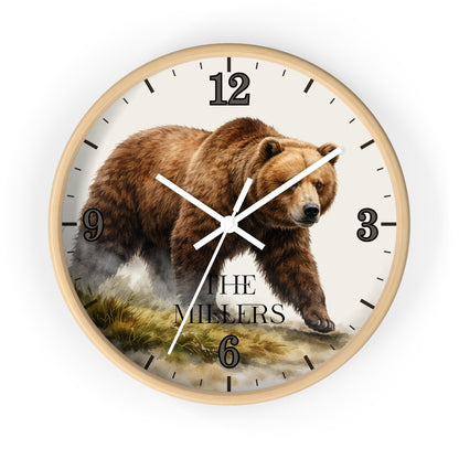 A round wall clock with a light wood frame and a printed design featuring a brown grizzly bear walking across a landscape. The clock face is off-white with black Arabic numerals and hands, and the words 'THE MILLERS' are printed near the bottom.