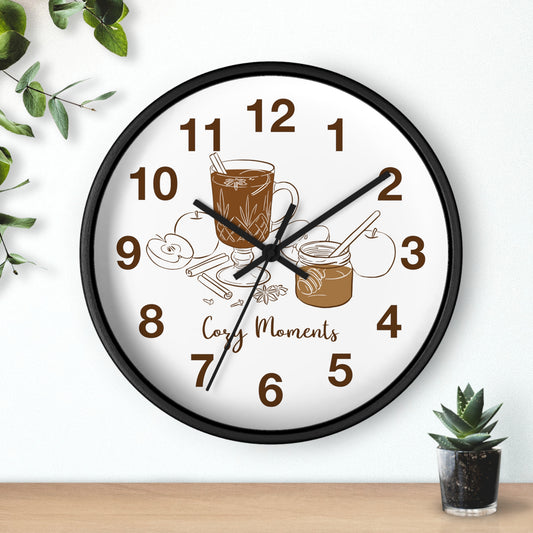 A round wall clock with a black frame and a white face, decorated with brown illustrations of a coffee mug, cookies, jam, and the words 'Cozy Moments'. The clock is mounted on a white wall above a wooden surface, with decorative green leaves and a small potted succulent nearby.