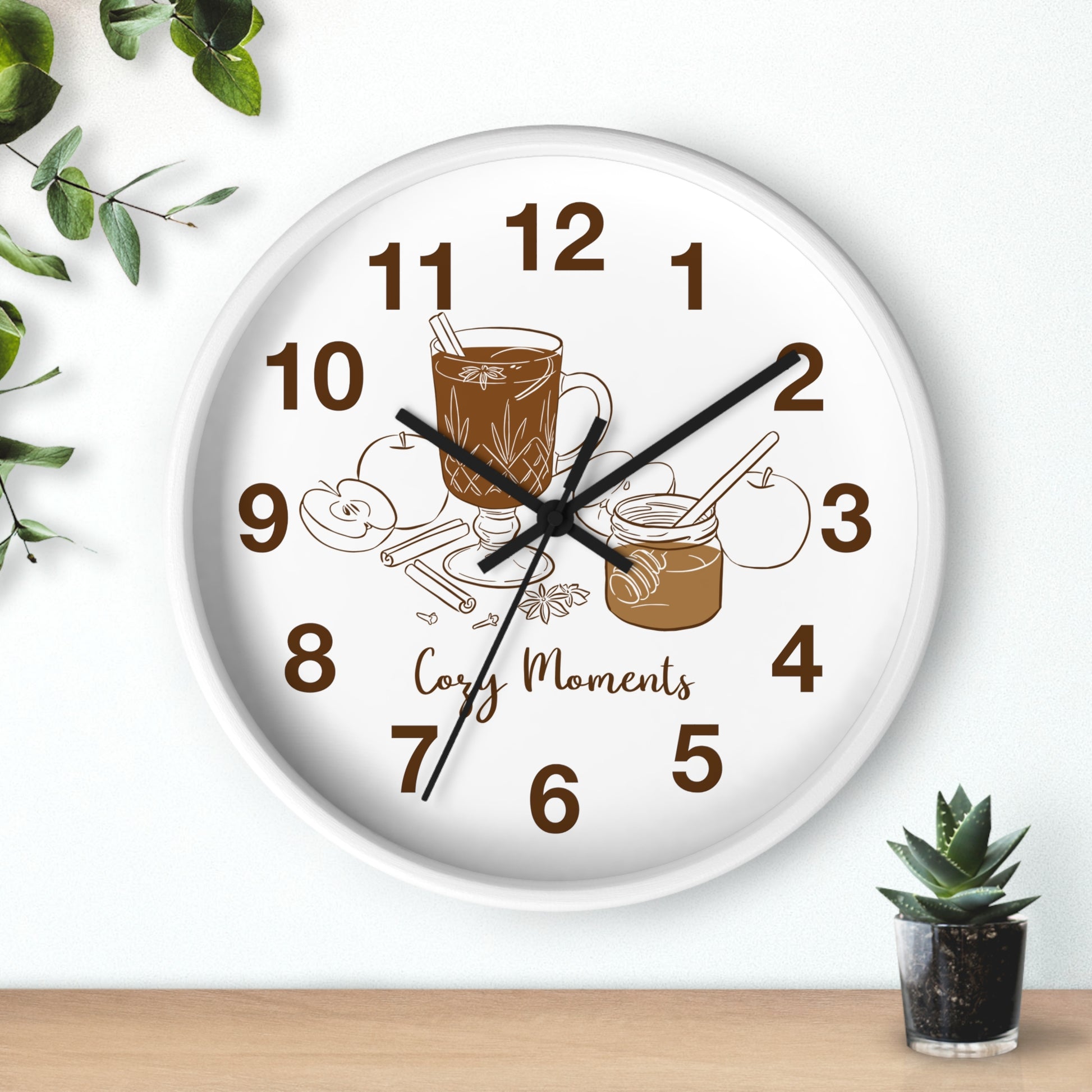 A round white wall clock with a warm-toned coffee-themed illustration and the words 'Cozy Moments' on the face. The clock is mounted on a white wall above a light wood surface, with decorative green leaves and a small potted succulent nearby.