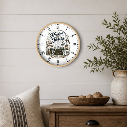 A round wall clock with a light wood frame hangs on a white shiplap wall above a wooden side table. The clock face is white with black Roman numerals and a rustic illustration featuring the words 'Hooked on Jesus' and 'Mark 7:14'.