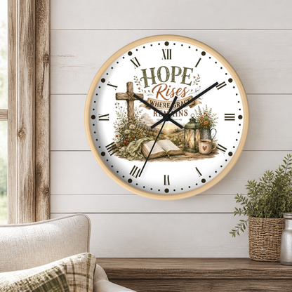 A round wall clock with a light wood frame hangs on a white shiplap wall above a wooden table. The clock face features a rustic Christian illustration with the words 'HOPE RISES WHERE GRACE REMAINS' and a cross, set in a styled home interior with a chair and a potted plant.