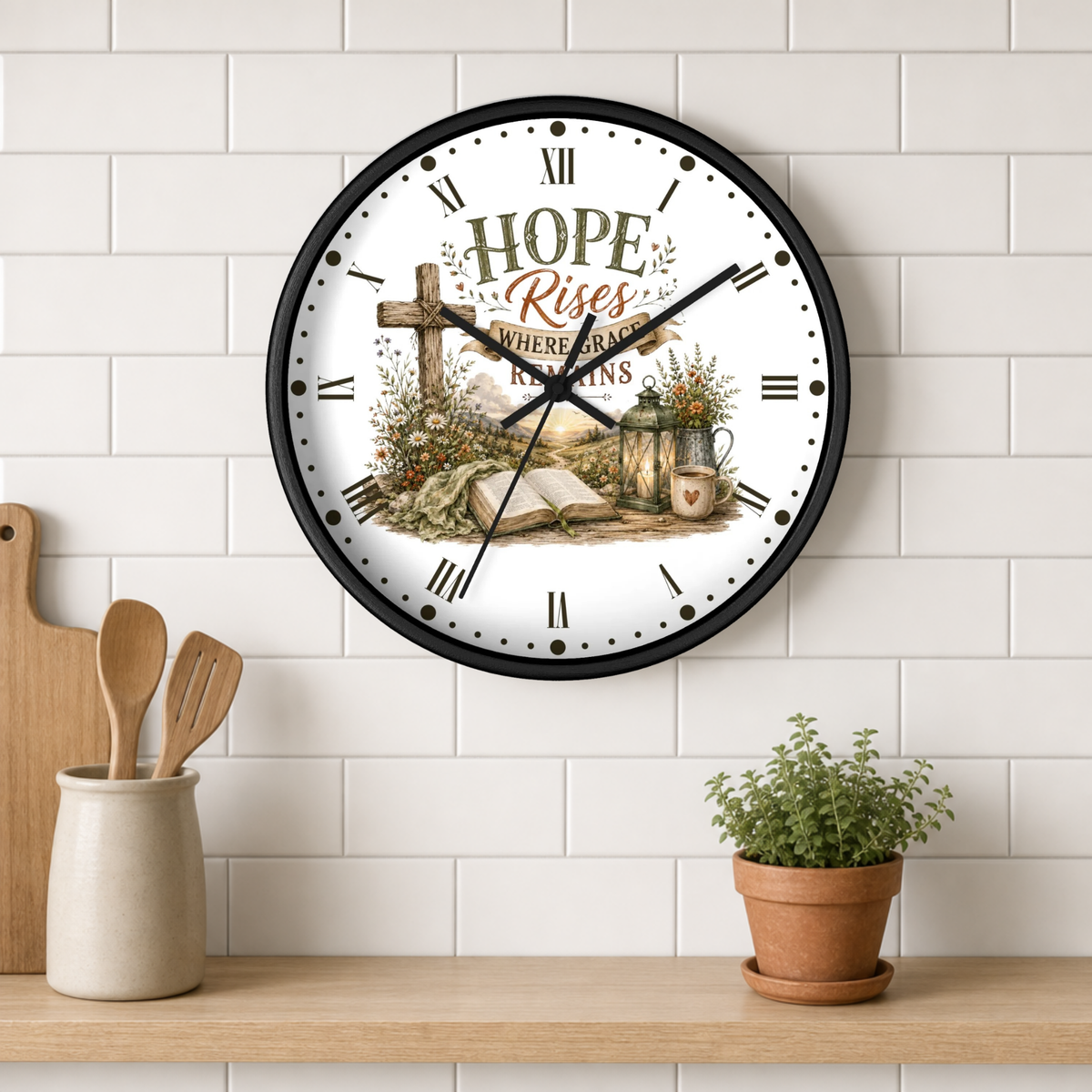 A round wall clock with a black frame and a white face featuring the words 'HOPE Rises WHERE GRACE REMAINS' over a rustic cross, flowers, and an open book. The clock is mounted on a white subway tile wall above a wooden shelf with kitchen utensils and a small potted plant.