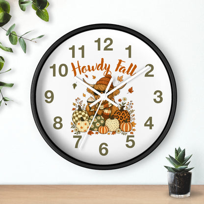A round wall clock with a black frame and a white face, decorated with autumn-themed graphics including pumpkins, leaves, and the words 'Howdy Fall' in orange script. The clock is shown hanging on a white wall above a wooden surface, with small green plants in the corners.