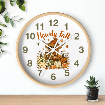 A round wall clock with a light wood frame and a white face, decorated with autumn-themed graphics including pumpkins, leaves, and the words 'Howdy Fall' in orange script. The clock is shown hanging on a white wall above a wooden surface, with a small potted succulent and green leaves as props.