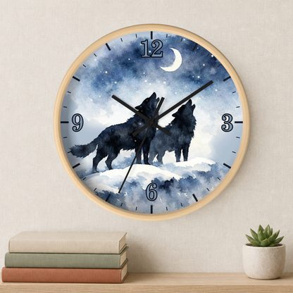 A round wall clock with a light wood frame hangs on a beige wall above a shelf with stacked books and a small potted succulent. The clock face features a watercolor-style night scene with two black howling wolves under a crescent moon and stars in blue, white, and gray tones.