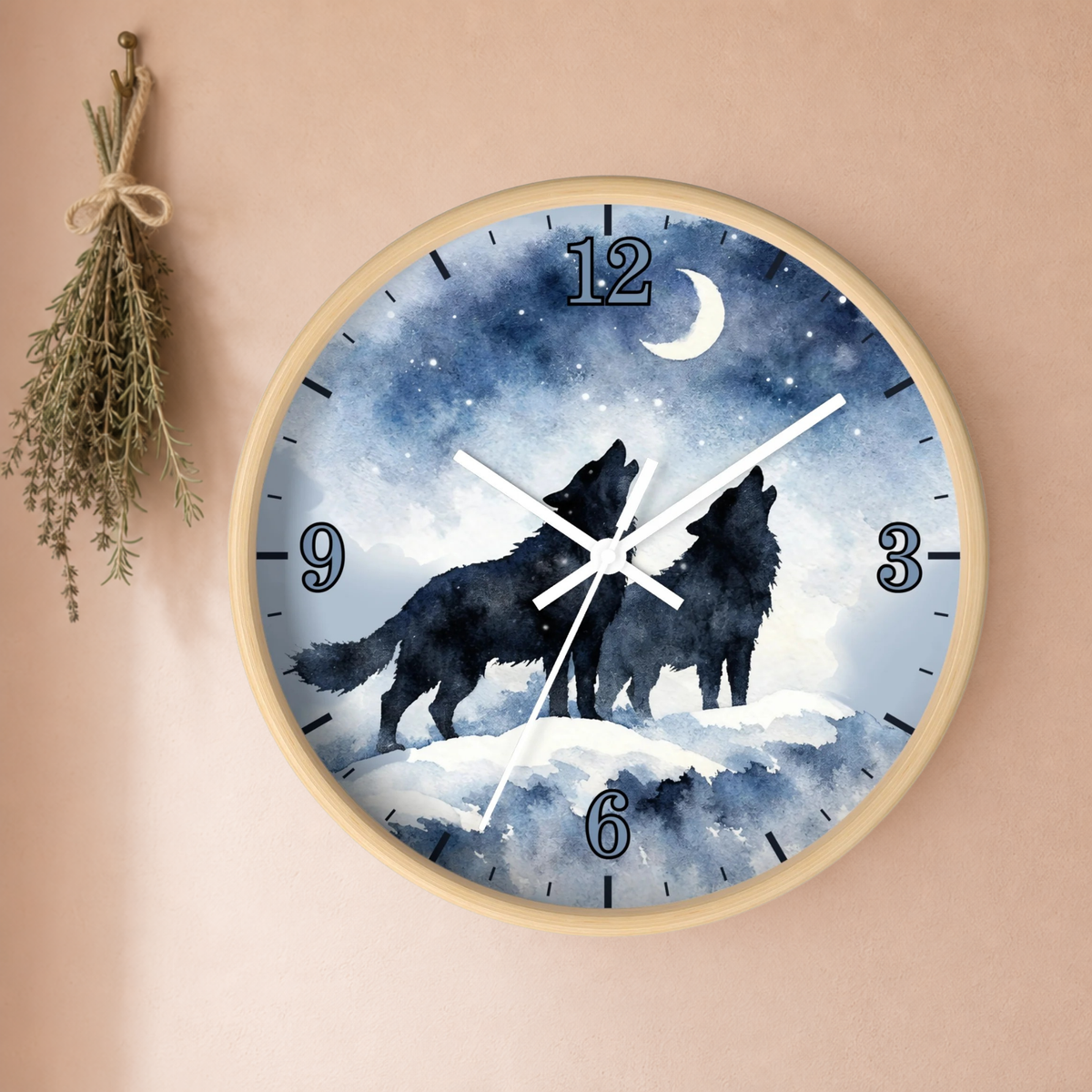 A round wall clock with a light wood frame, featuring a watercolor design of two black wolves howling at a snowy moonlit sky. The clock is mounted on a warm beige wall next to a hanging sprig of dried herbs.