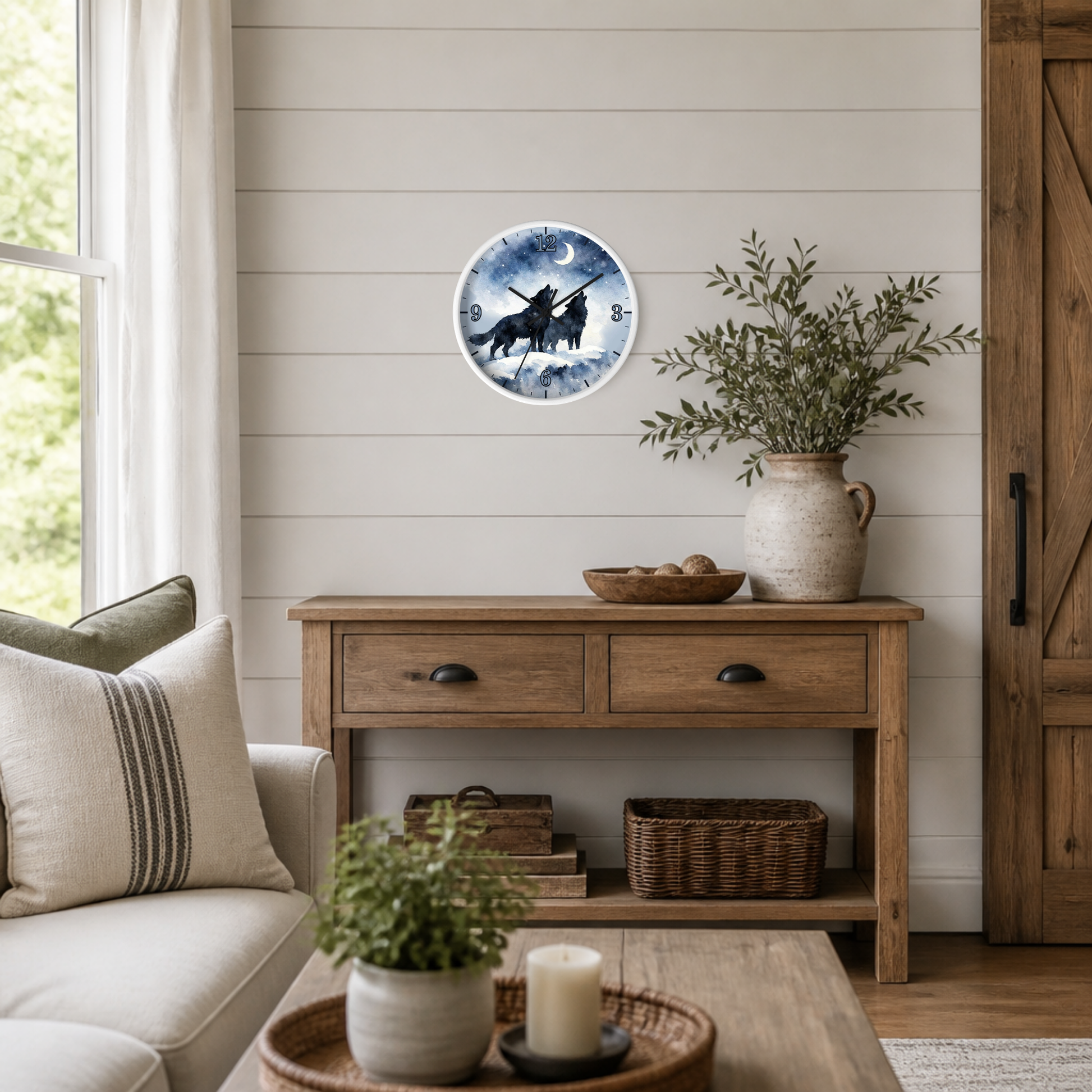 A round wall clock featuring a black silhouette of two wolves under a full moon and stars against a blue night sky, styled on a white shiplap wall above a rustic wooden console table in a cozy living room.