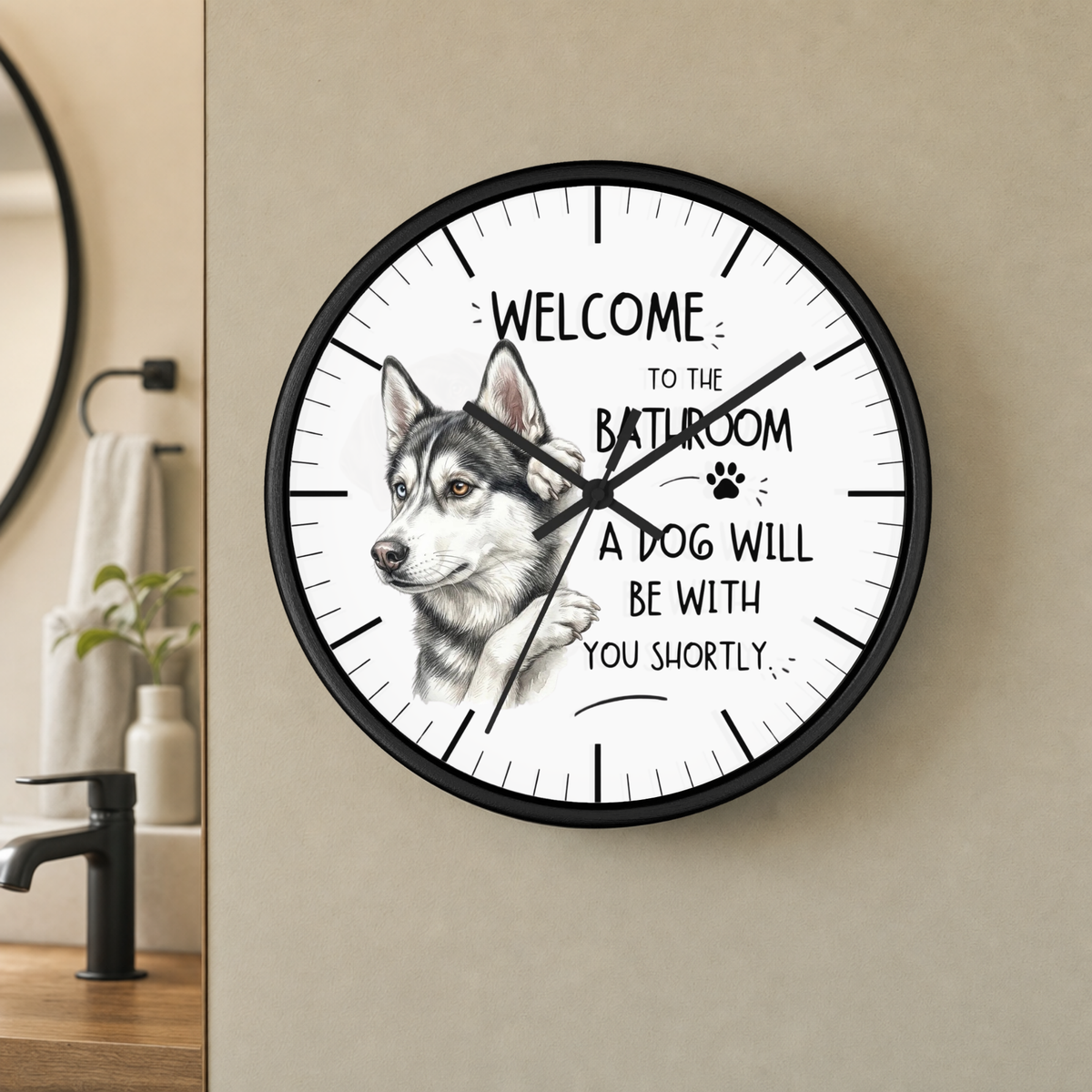 A round wall clock with a black frame and a white face, featuring a husky illustration and the text 'WELCOME TO THE BATHROOM A DOG WILL BE WITH YOU SHORTLY'. The clock is mounted on a beige wall in a bathroom setting.